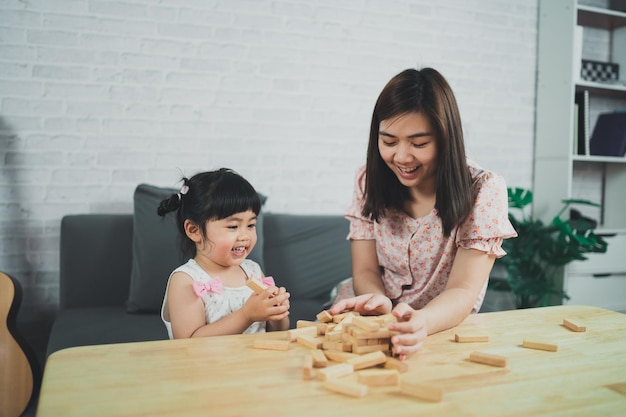 Concepto de juegos de jenga Hija y madre asiáticas sonriendo alegremente y riendo jugando juegos de jenga de madera sentados en un sofá en la sala de estar en casa actividades familiares de madre e hija jugando juegos felices