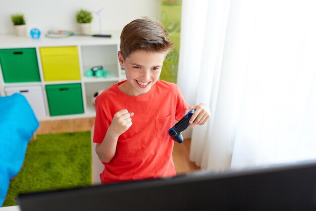 concepto de juegos, tecnología y personas - niño sonriente con gamepad y computadora jugando videojuegos y celebrando la victoria en casa