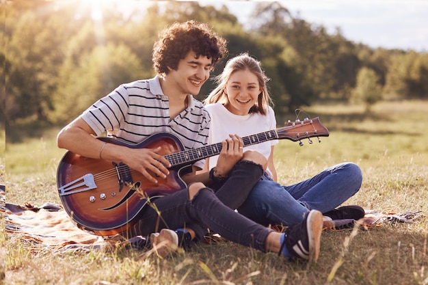 Concepto de juventud, citas y tiempo de recreación. La novia y el novio felices se sientan en la hierba verde, tocan la guitarra, disfrutan de un ambiente tranquilo