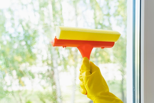 El concepto de lavado de ventanas. Una ama de casa con guantes amarillos lava la ventana con un raspador y una esponja