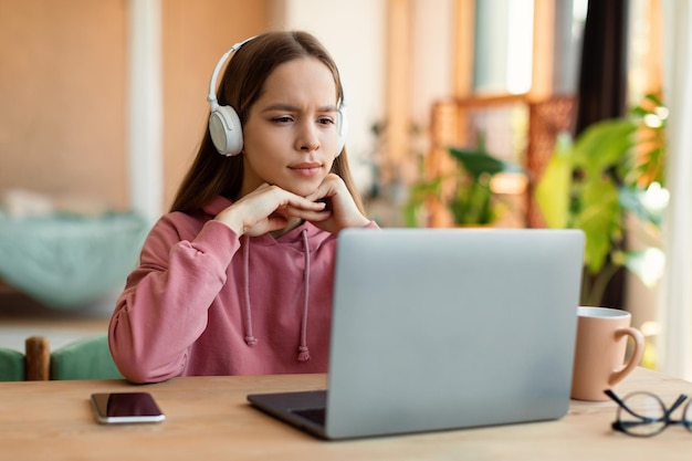 Concepto de lecciones en línea de educación Chica adolescente enfocada en auriculares mirando la pantalla del portátil sentada en la mesa en casa