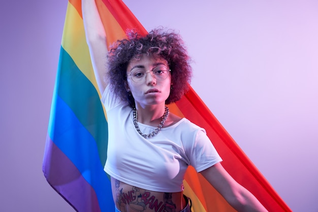 Concepto lgbtq. Chica caucásica positiva con pelo rizado afro sosteniendo la bandera del arco iris aislado en estudio