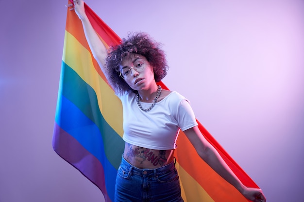 Concepto lgbtq. Chica caucásica positiva con pelo rizado afro sosteniendo la bandera del arco iris aislado