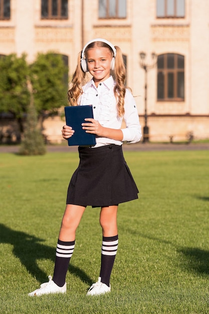 Concepto de libro de audio. Escuchar libro escolar. Tecnologías digitales para el aprendizaje. Elearning y métodos modernos. Niña linda colegiala mantenga libro y auriculares. El conocimiento se asimila mejor de esta manera.