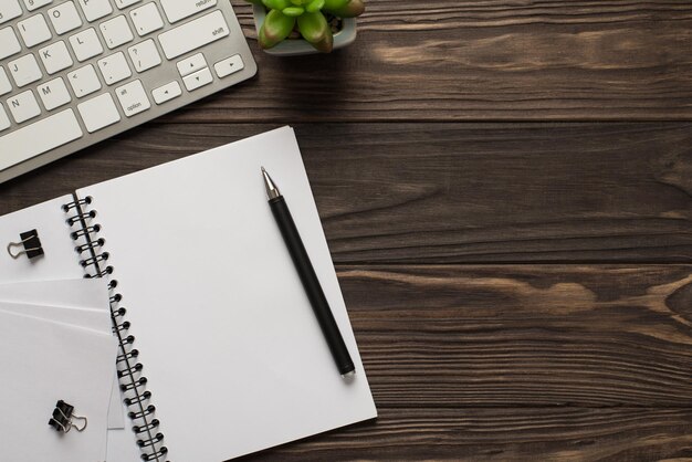 Concepto de lugar de trabajo empresarial. Endecha plana superior arriba ver foto de teclado blanco suculento y abierto libreta en blanco transparente con clips de encuadernación aislado mesa de madera oscura