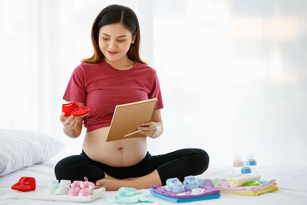 Concepto de madre feliz y saludable. Mujer embarazada asiática sentada en una cama sosteniendo un libro y un lápiz mientras sonríe y mira un lindo zapato de bebé rojo lleno de amor.