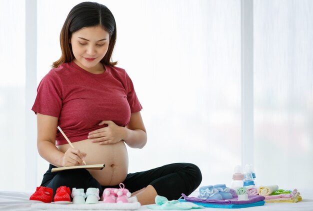 Concepto de madre feliz y saludable. Mujer embarazada asiática sentada en una cama sosteniendo un libro y un lápiz mientras sonríe y mira un lindo zapato de bebé rojo lleno de amor.