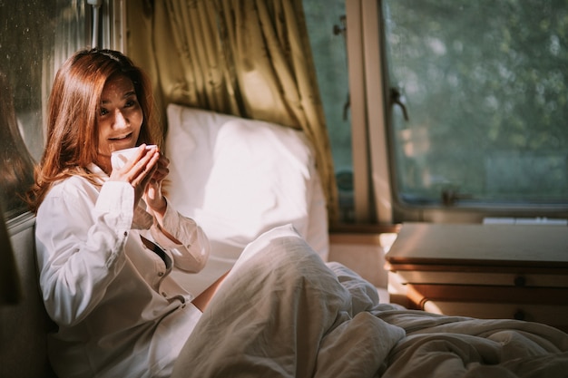 Concepto de mañana, comodidad, invierno y personas - cerca de mujer joven feliz con una taza de café o bebida de cacao en la cama