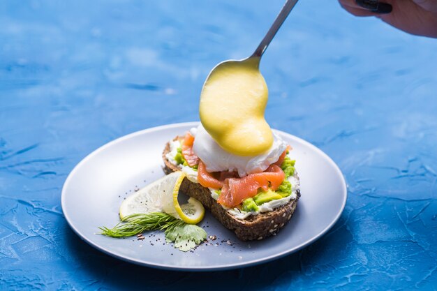 Concepto de mañana y desayuno. Preparación de sándwich con salmón, cuchara vierte salsa sobre él.