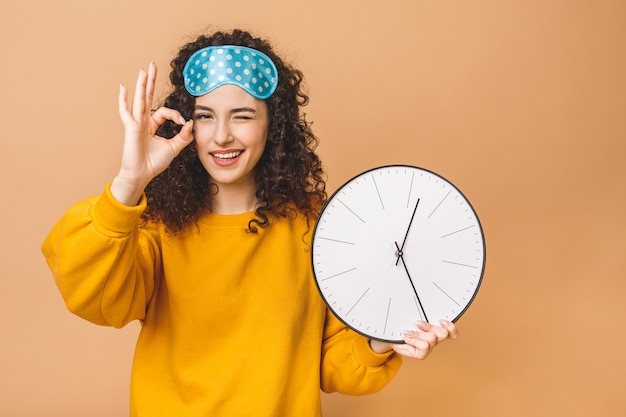 Foto concepto de mañana hermosa joven rizada posando sobre fondo beige con reloj y máscara para dormir. ok signo