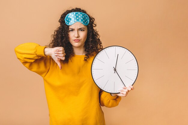 Foto concepto de mañana hermosa joven rizada posando sobre fondo beige con reloj y máscara para dormir.