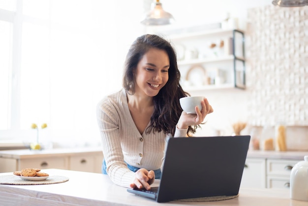 En el concepto de la mañana mujer feliz usando una computadora portátil y bebiendo café caliente sentado en la mesa en la cocina