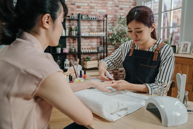 Concepto de manicura en proceso. dos clientes y manicuristas chinas asiáticas sentadas y sonriendo en el salón de spa. Esteticista recortando cutículas por clipper de clienta en taller de belleza moderno.