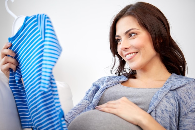 concepto de maternidad, embarazo, personas y ropa para niños - mujer feliz sosteniendo y mirando el traje azul de los bebés en casa