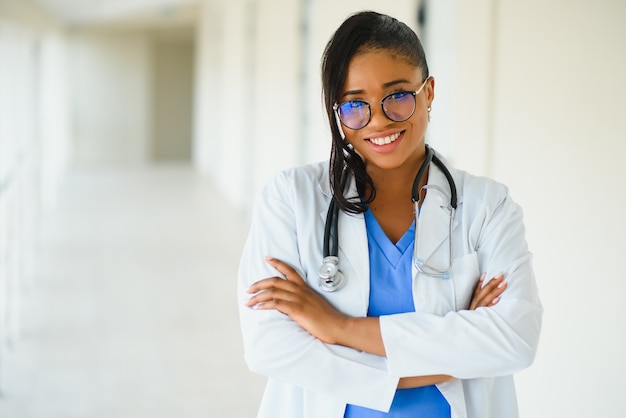 Concepto de medicina, profesión y cuidado de la salud - feliz sonriente doctora afroamericana en bata blanca con estetoscopio sobre fondo de hospital