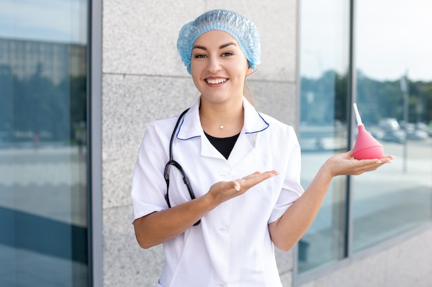 Concepto de medicina, profesión y personas: una enfermera europea sonriente con una bata blanca con un estetoscopio y un gorro médico desechable en la cabeza sosteniendo y apuntando a un enema al aire libre