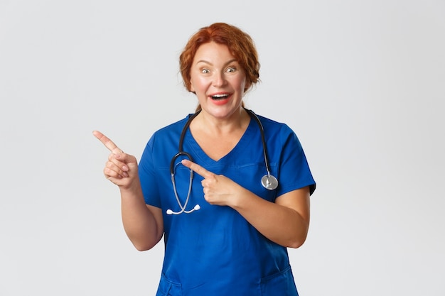 Concepto de medicina, salud y coronavirus. Enfermera pelirroja alegre emocionada, trabajador médico en matorrales apuntando hacia la esquina superior izquierda. Doctor mostrando oferta de promoción impresionante, sonriendo sorprendido.