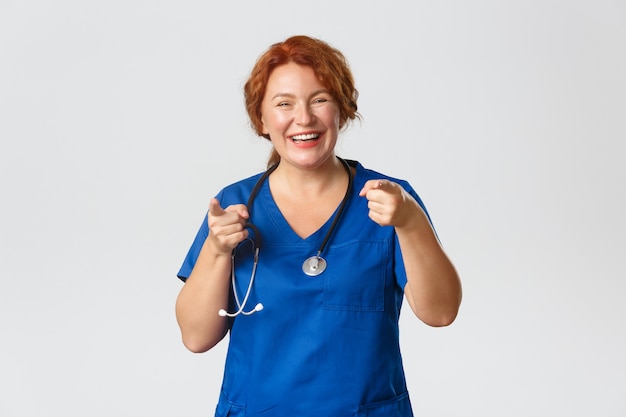 Concepto de medicina, salud y coronavirus. Trabajador médico pelirrojo alegre y entusiasta, doctora en matorrales apuntando con el dedo a la cámara y sonriendo, invitándolo, alabando la buena elección.