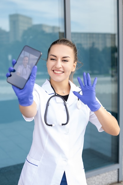 Concepto de medicina, salud, profesión y personas: una doctora europea sonriente con un estetoscopio en una bata médica y guantes de goma tomando selfies en su teléfono y saludando a la cámara