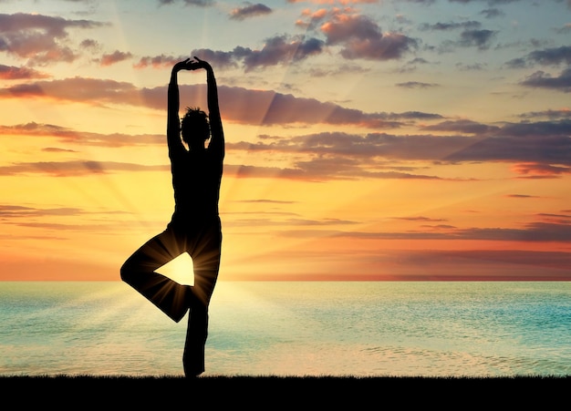 Concepto de meditación y relajación. Silueta de una niña practicando clase de yoga sobre un fondo de puesta de sol del mar