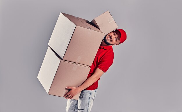 Concepto de mensajería y mensajería. Hacer muecas chico guapo está tratando de mantener una gran pila de cajas de cartón en su mano, aislado sobre fondo gris.