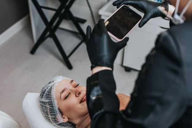 Concepto de microblading de cejas. Cosmetóloga preparando a una mujer joven para el procedimiento de maquillaje permanente de cejas y tomando una foto de sus cejas con un teléfono inteligente.
