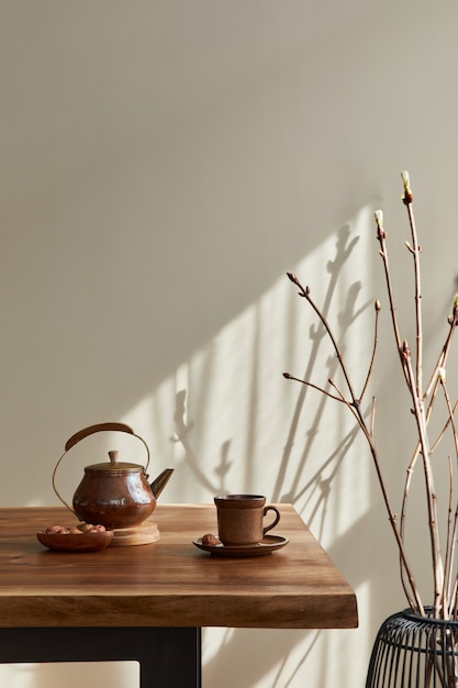 Concepto minimalista del interior del comedor con mesa familiar de madera, taza de café, jarra de té, vajilla, pared beige y accesorios personales. Copie el espacio ..