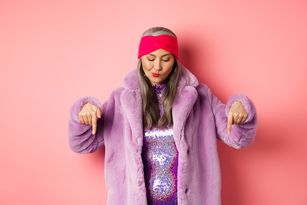 Foto concepto de moda y compras. funky mujer mayor asiática en abrigo de piel sintética de moda y vestido de discoteca, mirando y apuntando hacia abajo, de pie sobre fondo rosa.