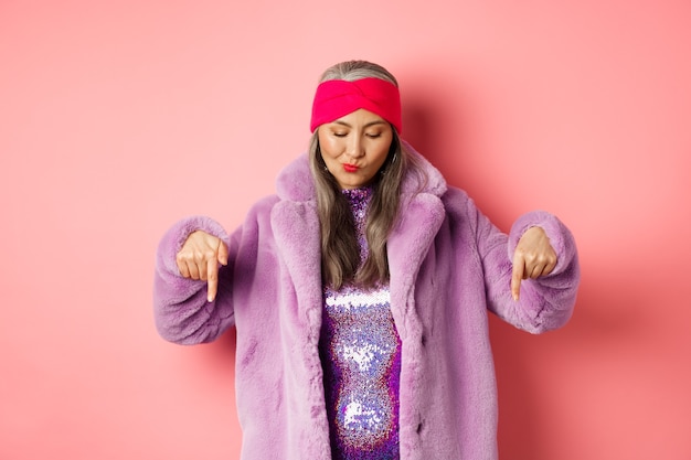 Concepto de moda y compras. Funky mujer mayor asiática en abrigo de piel sintética de moda y vestido de discoteca, mirando y apuntando hacia abajo, de pie sobre fondo rosa.