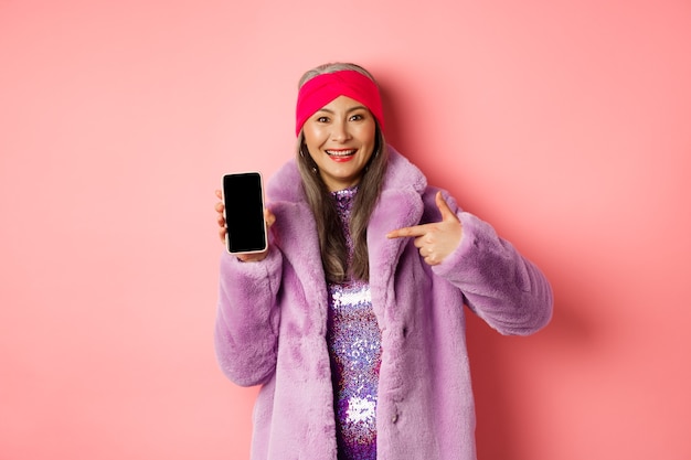 Concepto de moda y compras en línea. Elegante dama madura asiática que muestra la pantalla del teléfono inteligente en blanco, apuntando al teléfono y sonriendo, fondo rosa