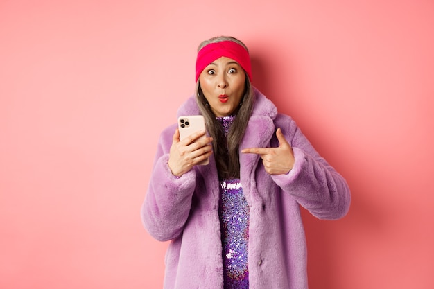 Concepto de moda y compras en línea. Feliz abuela asiática mirando una promoción impresionante, señalando con el dedo al teléfono inteligente y mirando asombrado, fondo rosa