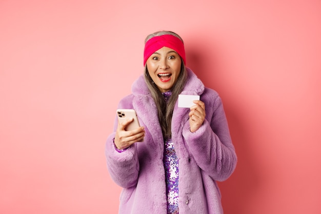 Concepto de moda y compras en línea. Feliz mujer asiática revisando promociones de internet, con tarjeta de crédito y teléfono inteligente, fondo rosa.