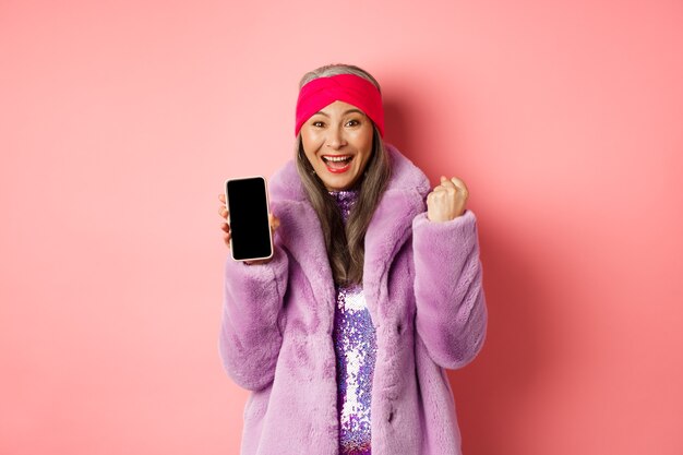 Concepto de moda y compras en línea. Feliz mujer mayor asiática ganando el premio en internet, mostrando la pantalla del teléfono móvil en blanco y haciendo bomba de puño, grito de alegría, de pie sobre fondo rosa.