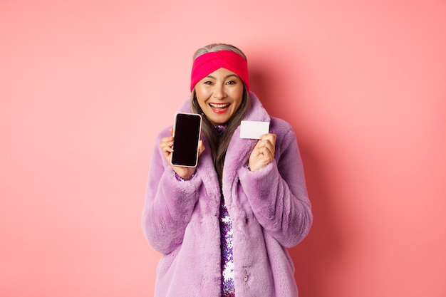 Concepto de moda y compras en línea. Hermosa mujer madura asiática mostrando tarjeta de crédito de plástico y pantalla de teléfono en blanco, sonriendo feliz, fondo rosa