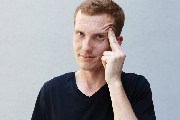 Concepto de moda, estilo, emociones y personas - Close up retrato de una pelirroja de un hermoso chico varonil con pecas en una camiseta negra de fondo gris. pensativamente sostiene una mano por la sien