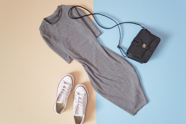 Concepto de moda. Estilo urbano para mujer. Vestido gris, zapatillas blancas y bolso negro sobre fondo azul suave, beige.