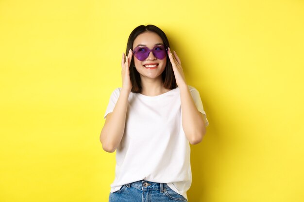 Concepto de moda y estilo de vida. Elegante mujer asiática probando nuevas gafas de sol en forma de corazón, yendo de vacaciones, sonriendo feliz a la cámara, de pie sobre fondo amarillo.