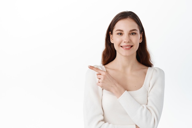 Concepto de moda y estilo de vida de primavera Mujer joven con estilo segura en traje apuntando con el dedo a la izquierda y mirando algo con una sonrisa complacida encontró un gran producto de fondo blanco