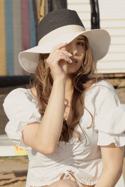 Concepto de moda de vacaciones de verano - mujer de bronceado con sombrero para el sol en la playa con una blusa blanca y bikini negro. Chica con sombrero sentada en la hamaca. Viajes y vacaciones de verano