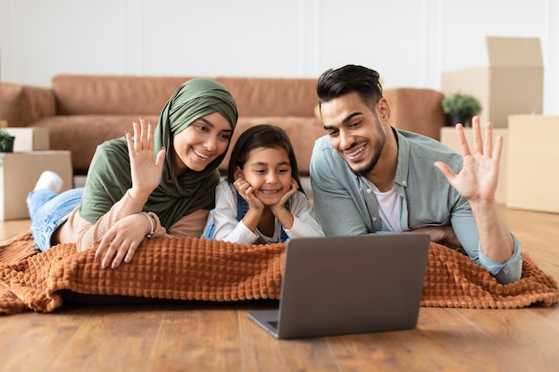 Concepto moderno de comunicación remota. Retrato de familia árabe positiva haciendo videollamadas en línea con amigos o abuelos usando una computadora portátil, acostado en la alfombra del piso, saludando con la mano mirando la pantalla