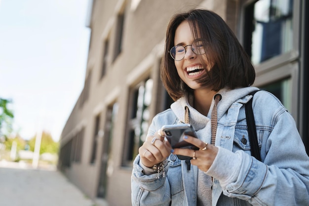 Concepto moderno de generación y tecnología Retrato al aire libre de una chica hipster despreocupada con gafas de pie en la calle riéndose a carcajadas con los ojos cerrados sosteniendo un mensaje divertido de lectura de teléfono móvil