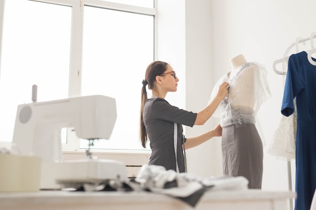 Concepto de modista, sastre, moda y sala de exposición - Vista lateral del diseñador de moda femenina midiendo materiales en maniquí en la oficina.