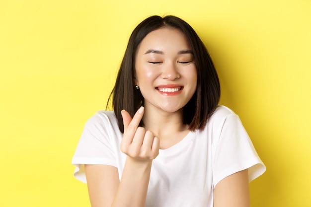 Concepto de mujer y día de San Valentín. Cerca de una chica bastante asiática en camiseta blanca, sonriendo y mostrando el corazón del dedo, te amo gesto, de pie sobre fondo amarillo.