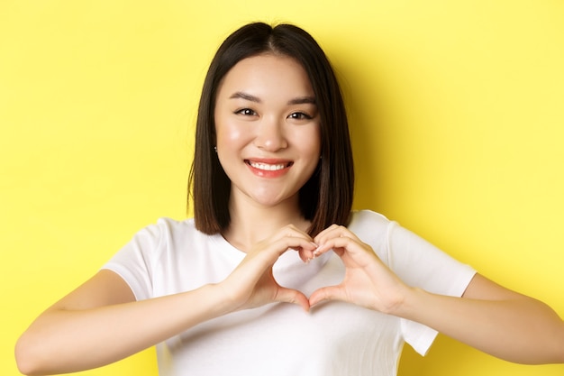 Concepto de mujer y día de San Valentín. Cerca de una chica bastante asiática en camiseta blanca, sonriendo y mostrando el corazón, te amo gesto, de pie sobre fondo amarillo.