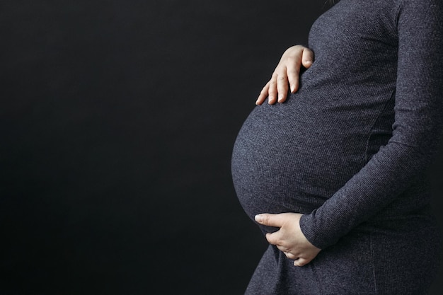 El concepto de una mujer embarazada Una mujer embarazada con un vestido sostiene su estómago con las manos al lado sobre un fondo negro hay espacio libre para texto