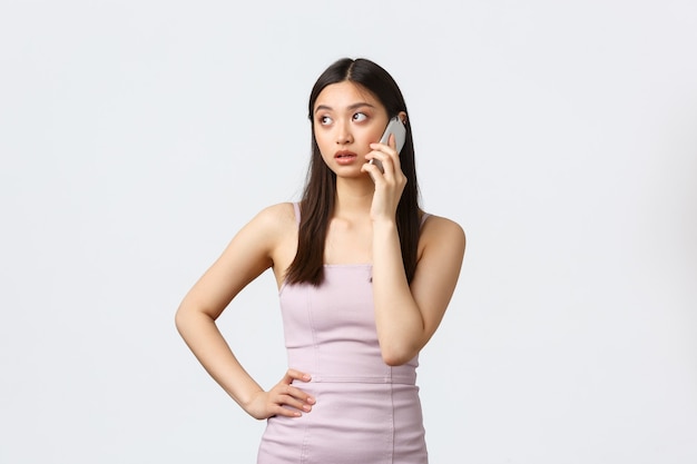 Concepto de mujer, fiesta y vacaciones de lujo. Mujer asiática impaciente en traje de noche, mirando a otro lado pensativo, llamando, teniendo una llamada telefónica