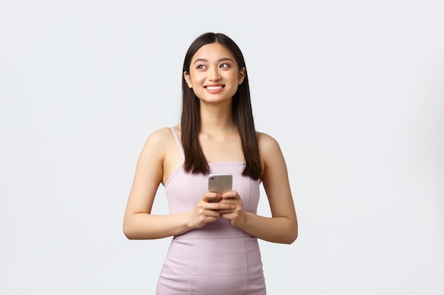 Concepto de mujer, fiesta y vacaciones de lujo. Satisfecho, feliz, soñadora chica asiática en vestido de noche mirando hacia la esquina superior izquierda encantada, usando teléfono móvil