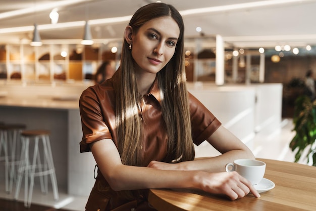 Concepto de mujeres empoderadas Atractiva y exitosa mujer de negocios joven sentada en un café bebiendo una taza de café inclinando la cabeza y mirando la cámara con interés sonriendo seductora