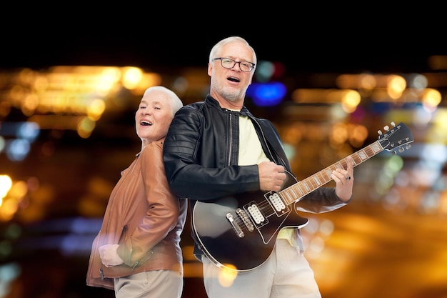 concepto de música, edad y gente - feliz pareja de ancianos con guitarra eléctrica cantando sobre luces nocturnas de fondo
