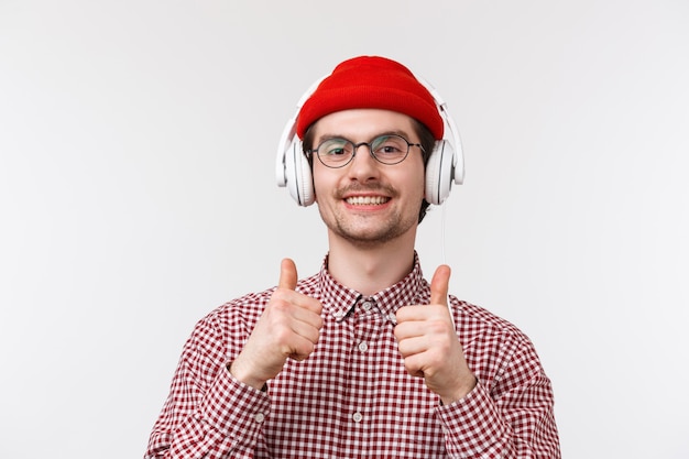 Concepto de música, tecnología y personas. Close-up retrato de satisfecho satisfecho joven caucásico hipster barbudo hombre con gafas y gorro, mostrar el pulgar hacia arriba como escuchar música en los auriculares